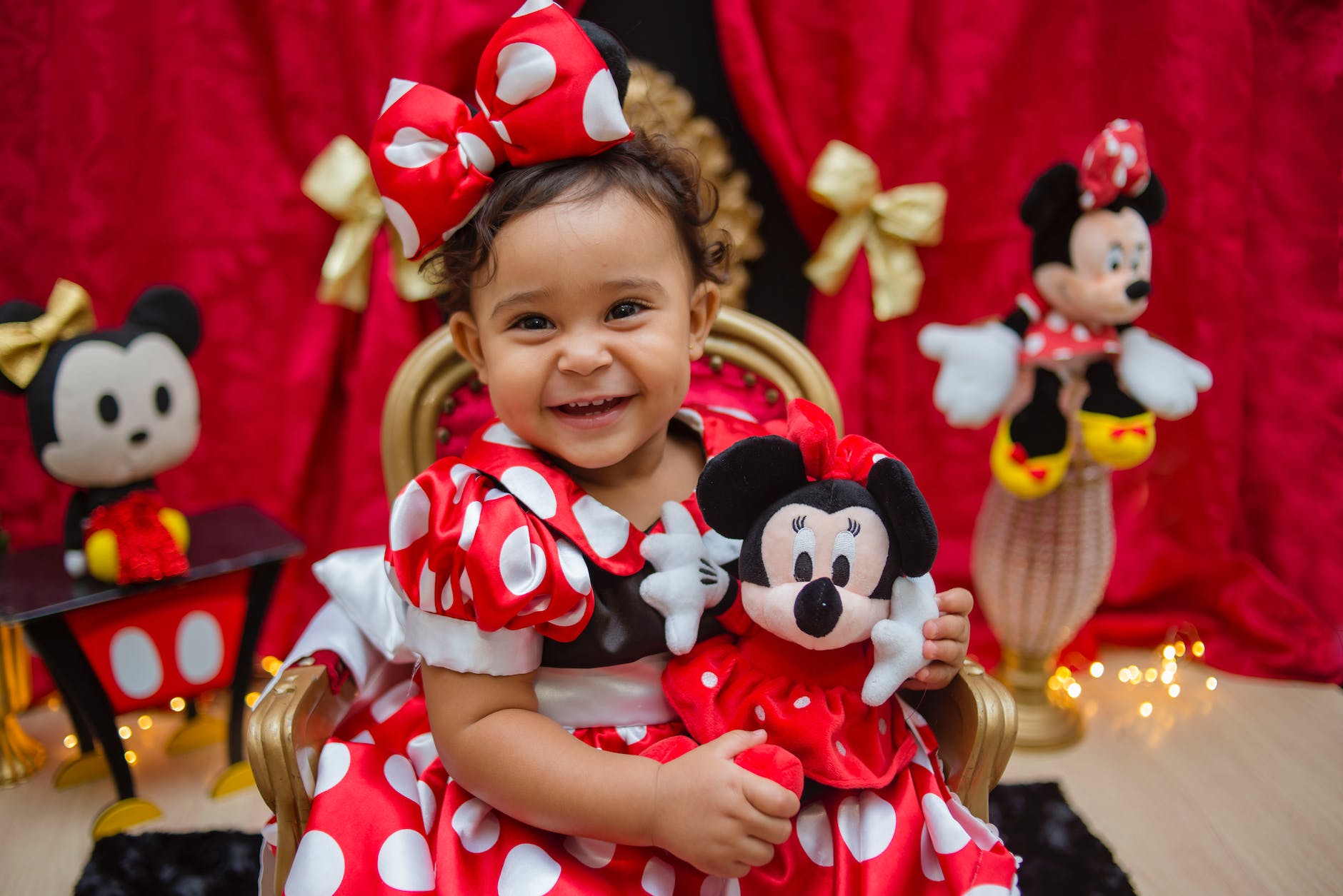 a cute girl in red polka dots dress sitting while holding a mickey mouse toy
